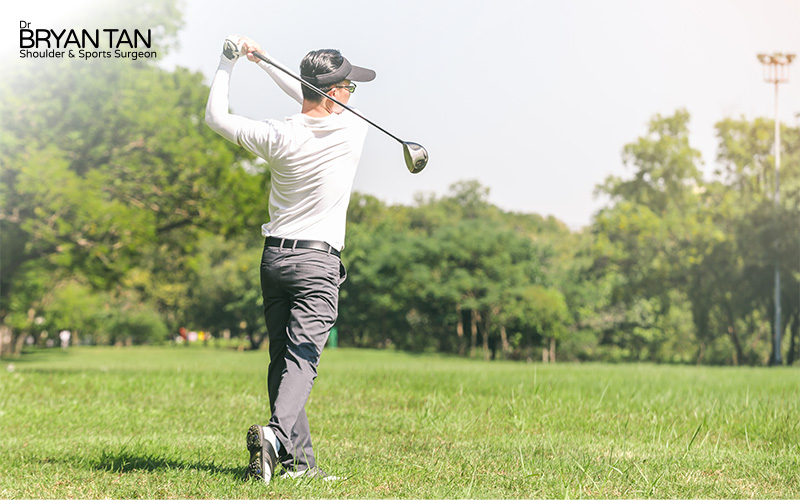 Golfer mid-swing on a sunny course, with lush trees and clear sky in the background.