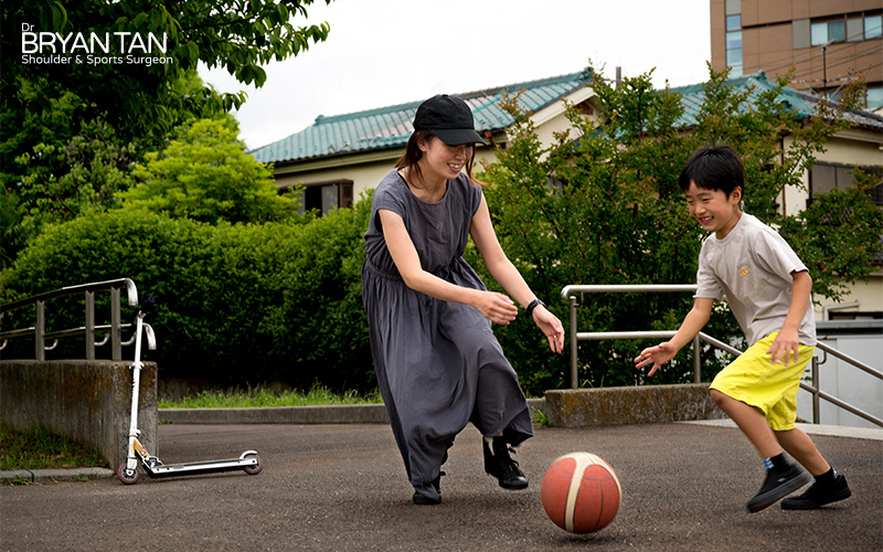 Woman and child playing basketball outdoors near a scooter and residential buildings.
